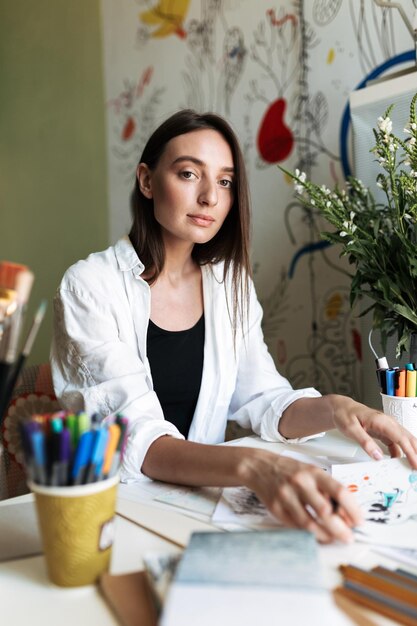 Young pensive painter sitting at the desk with pictures thoughtfully looking in camera while drawing at home