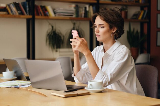 노트북과 커피 한 잔을 들고 탁자에 앉아 있는 수심에 찬 젊은 여성이 현대 사무실에서 휴대폰을 사용하면서 신중하게
