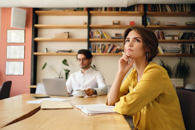Молодая задумчивая женщина сидит за столом с блокнотом и мечтательно смотрит в сторону, работая с коллегой на заднем плане в современном офисе