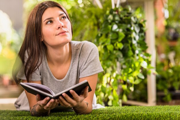 Young pensive woman with book lying on lawn