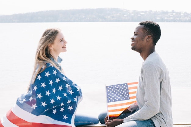 Foto gratuita giovani che si siedono sulla riva del mare e che sorridono insieme tenendo la bandiera americana