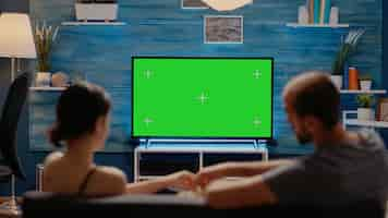 Free photo young people watching television with green screen display sitting in living room. caucasian man and woman with chroma key or copy space for isolated template and mockup template