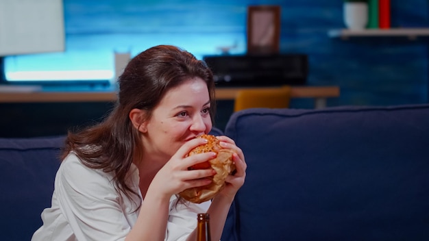 Foto gratuita giovane che ride alla tv mentre mangia hamburger e beve birra dalla bottiglia sul divano. donna che guarda la commedia e si gode un pasto da asporto fast food, seduta in soggiorno divertendosi