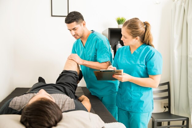 Young physiotherapist examining customer's leg while colleague noting in clipboard at hospital