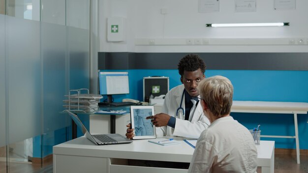 Young physiotherapist showing tablet of bones injury to senior patient with osteoporosis at desk in doctors office. Elder woman looking at virtual vertebrae for medical examination