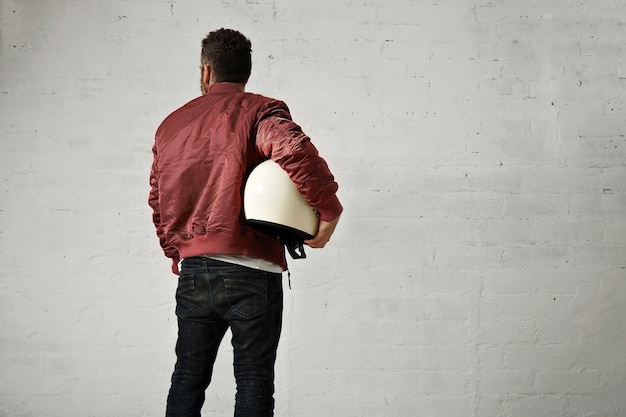 Free photo young pilot in jeans, shiny nylon burgundy bomber jacket and a round white helmet under his arm shot from the back against white wall background