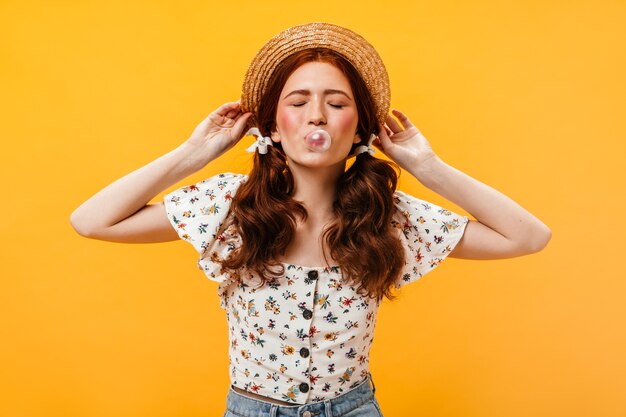 Young pink-cheeked woman with ponytails chews cud and puts on straw hat on orange background.