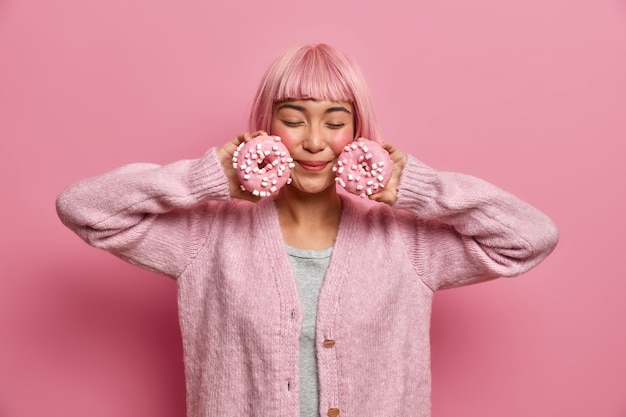 Free photo young pink haired woman enjoys taste of delicious donuts, poses with eyes closed, holds sweeties sprinkled doughnuts near face, wears warm sweater,