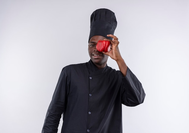 Young pleased afro-american cook in chef uniform closes eye with red pepper on white  with copy space