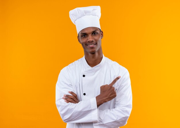 요리사 제복을 입은 젊은 만족 아프리카 계 미국인 요리사가 팔을 교차하고 주황색 벽에 고립 된 측면을 가리 킵니다.
