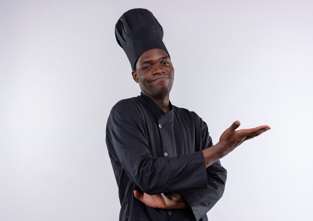Young pleased afro-american cook in chef uniform crosses arms and points at side with hand on white  with copy space