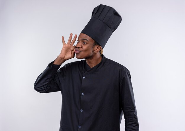 Молодой довольный афро-американский повар в униформе шеф-повара жестикулирует вкусный вкусный знак рукой на белой стене
