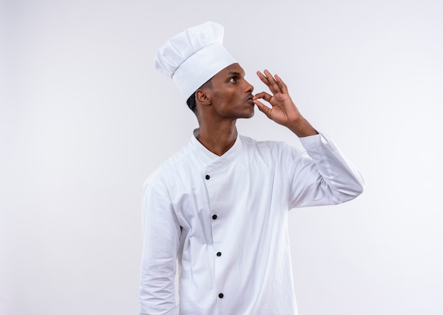 Young pleased afro-american cook in chef uniform gestures tasty delicious with hand isolated on white wall