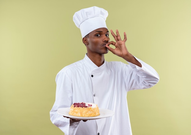 Young pleased afro-american cook in chef uniform holds cake on plate and gestures tasty deligious with hand isolated on green wall
