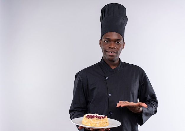 Young pleased afro-american cook in chef uniform holds cake on plate and points with hand on white  with copy space