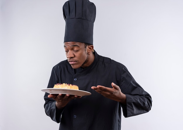 Young pleased afro-american cook in chef uniform holds cake on plate and pretends to smell isolated on white wall 