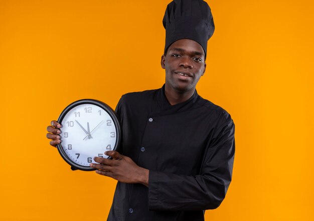 シェフの制服を着た若い喜んでいるアフリカ系アメリカ人の料理人は時計を保持し、コピースペースでオレンジ色のカメラを見ます