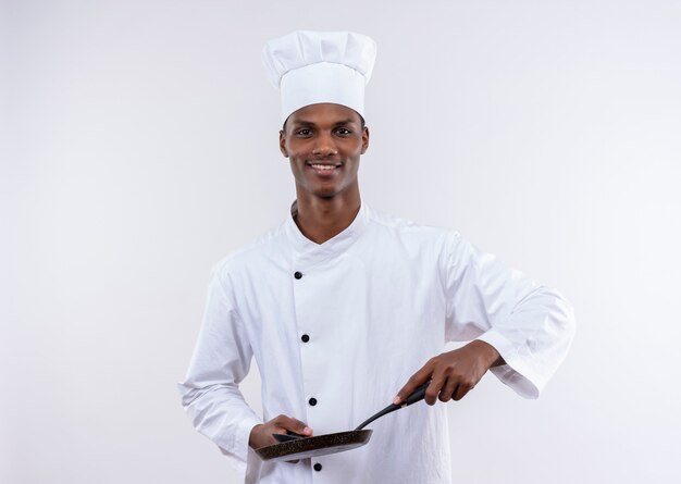 Young pleased afro-american cook in chef uniform holds frying pan and spoon on isolated white wall
