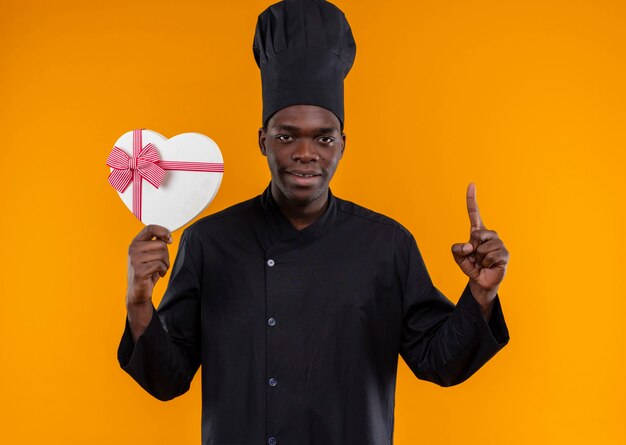 Young pleased afro-american cook in chef uniform holds heart shape box and points up on orange  with copy space