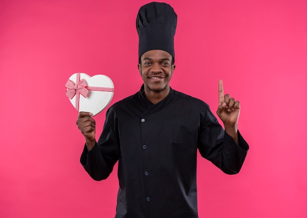 요리사 제복을 입은 젊은 만족 아프리카 계 미국인 요리사는 심장 모양 상자를 보유하고 분홍색 벽에 고립 된 엄지 손가락