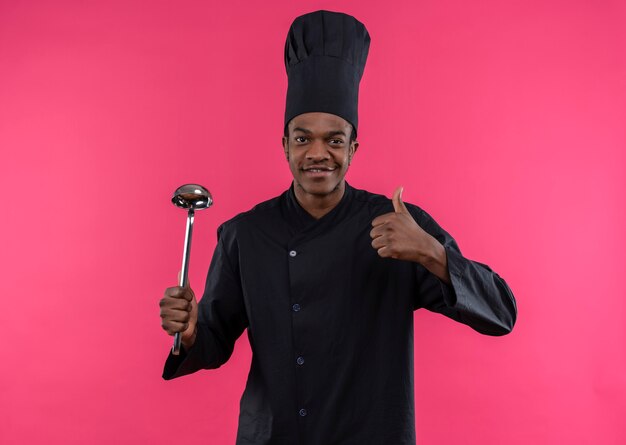 Young pleased afro-american cook in chef uniform holds ladle and thumbs up isolated on pink wall
