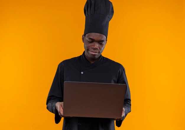 Il giovane cuoco afroamericano soddisfatto in uniforme del cuoco unico tiene ed esamina il taccuino sull'arancia con lo spazio della copia