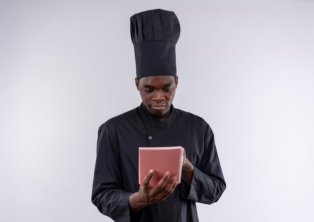 Young pleased afro-american cook in chef uniform holds and looks at notebook on white  with copy space