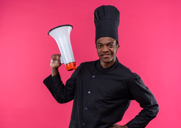 Young pleased afro-american cook in chef uniform holds loud speaker isolated on pink wall 