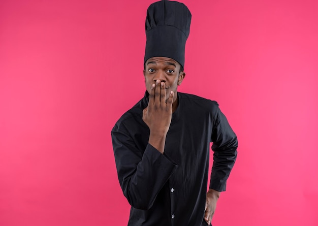 Young pleased afro-american cook in chef uniform holds mouth with hand isolated on pink wall 