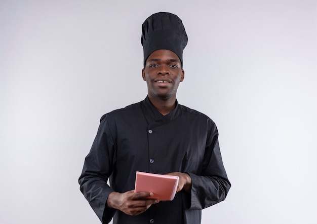 Young pleased afro-american cook in chef uniform holds and points at notebook on white  with copy space