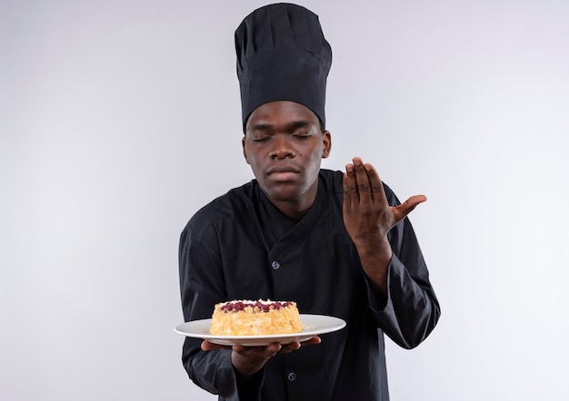 Young pleased afro-american cook in chef uniform holds and pretends to smell cake on plate on white  with copy space