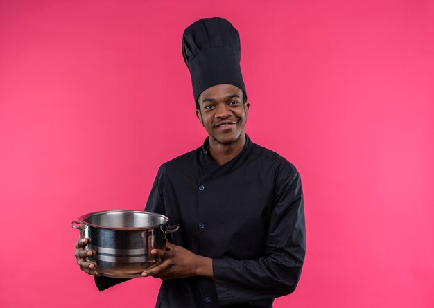 Young pleased afro-american cook in chef uniform holds saucepan isolated on pink wall 