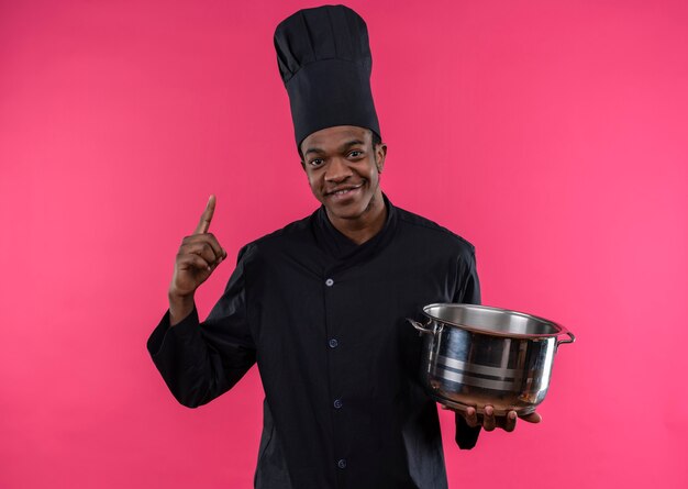 Young pleased afro-american cook in chef uniform holds saucepan and points up isolated on pink wall 