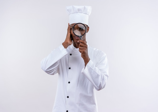 Young pleased afro-american cook in chef uniform looks through magnifying glass or loupe isolated on white wall