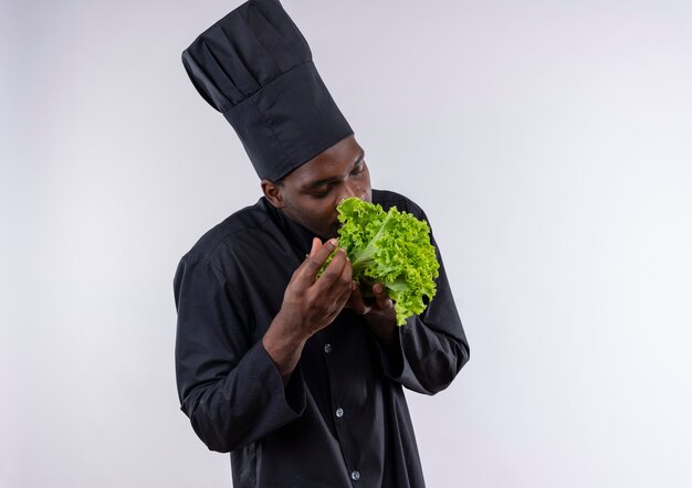 Young pleased afro-american cook in chef uniform pretends to smell salad on white  with copy space