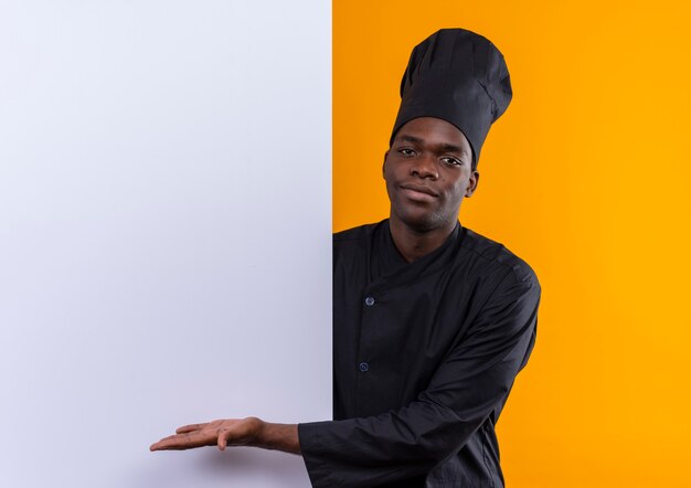Молодой довольный афро-американский повар в униформе шеф-повара стоит позади и указывает на белую стену на оранжевом фоне с копией пространства