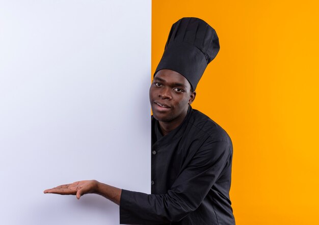 Young pleased afro-american cook in chef uniform stands behind and points with hand at white wall on orange  with copy space