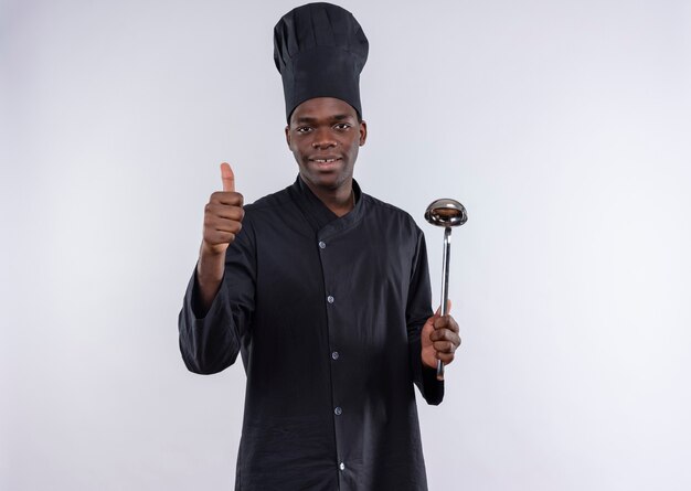 Young pleased afro-american cook in chef uniform thumbs up blinks with eye and holds ladle on white  with copy space