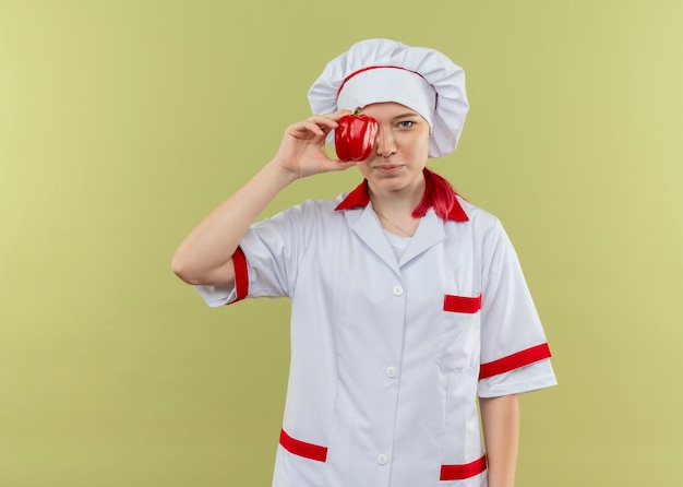 Young pleased blonde female chef in chef uniform closes eye with red pepper isolated on green wall