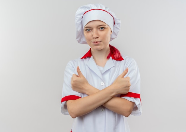 Young pleased blonde female chef in chef uniform crosses arms and thumbs up with both hands isolated on white wall