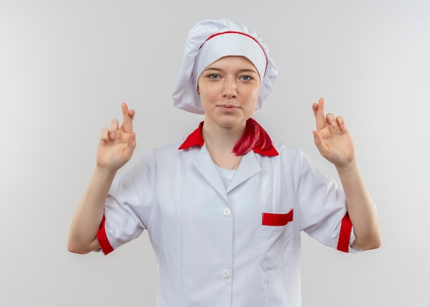 Young pleased blonde female chef in chef uniform crosses fingers isolated on white wall