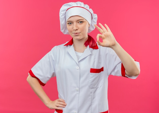 Foto gratuita giovane chef femmina bionda felice in uniforme del cuoco unico gesti il segno giusto della mano isolato sulla parete rosa