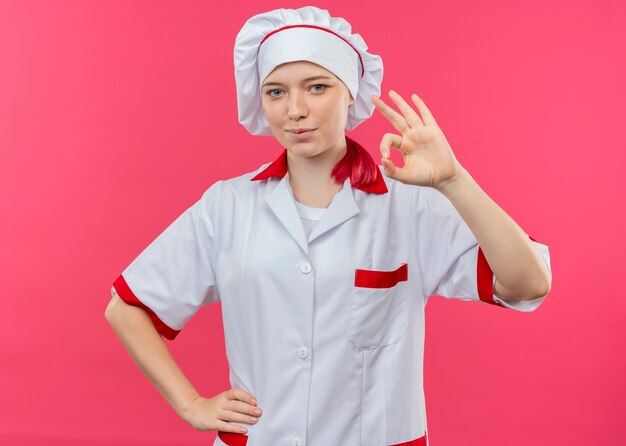 Young pleased blonde female chef in chef uniform gestures ok hand sign isolated on pink wall