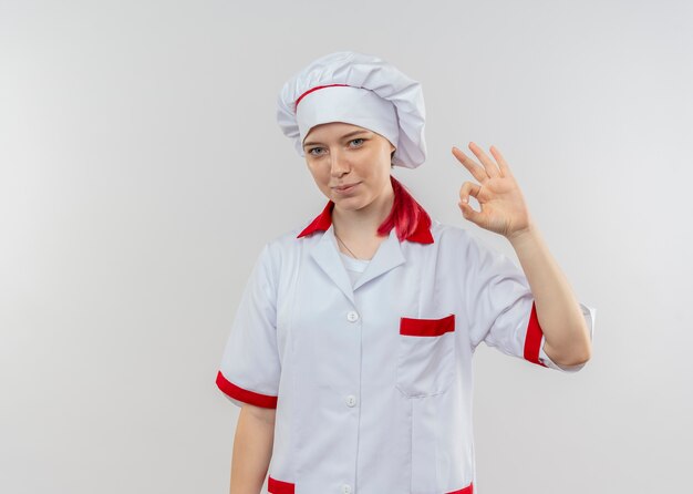 Молодая довольная блондинка-шеф-повар в униформе шеф-повара жестикулирует знак рукой на белой стене