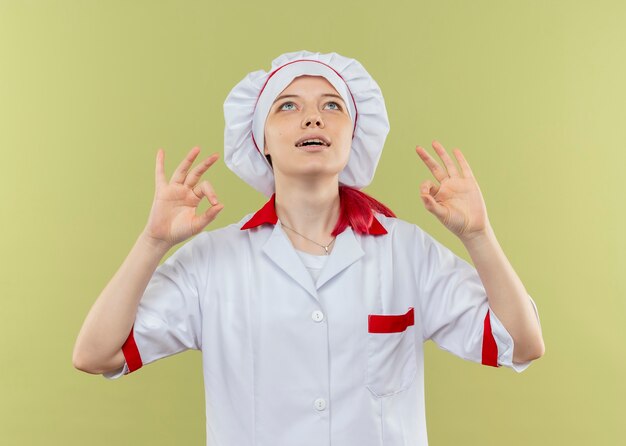 Young pleased blonde female chef in chef uniform gestures ok hand sign with hands and looks up isolated on green wall