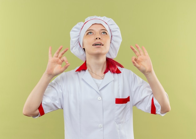 Young pleased blonde female chef in chef uniform gestures ok hand sign with hands and looks up isolated on green wall