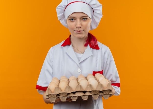 Young pleased blonde female chef in chef uniform holds batch of eggs and looks isolated on orange wall