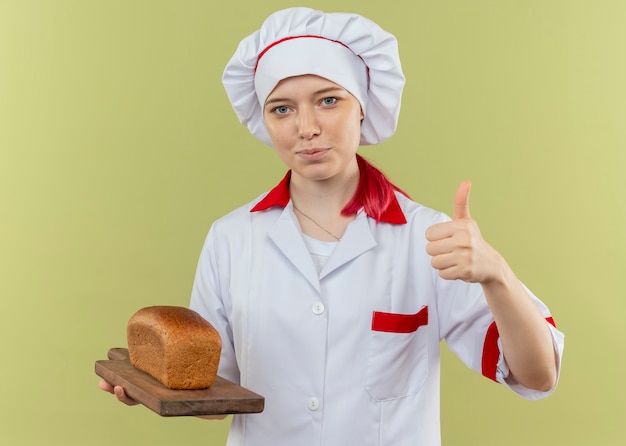 요리사 유니폼에 젊은 기쁘게 금발 여성 요리사는 커팅 보드에 빵을 보유하고 녹색 벽에 고립 엄지 손가락