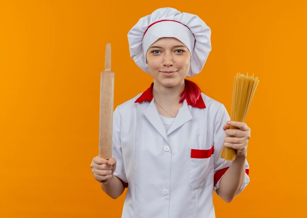 Young pleased blonde female chef in chef uniform holds bunch of spaghetti and rolling pin isolated on orange wall