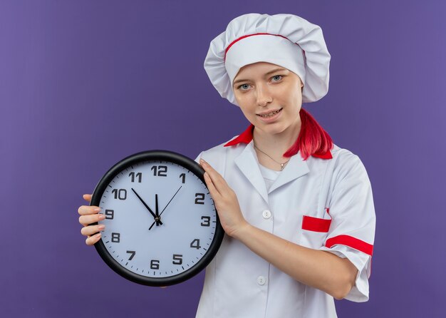 Young pleased blonde female chef in chef uniform holds clock and looks isolated on violet wall